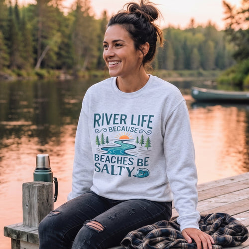 A woman sitting on a wooden dock by a lake at sunset, wearing a light gray sweatshirt with a blue and green graphic that read