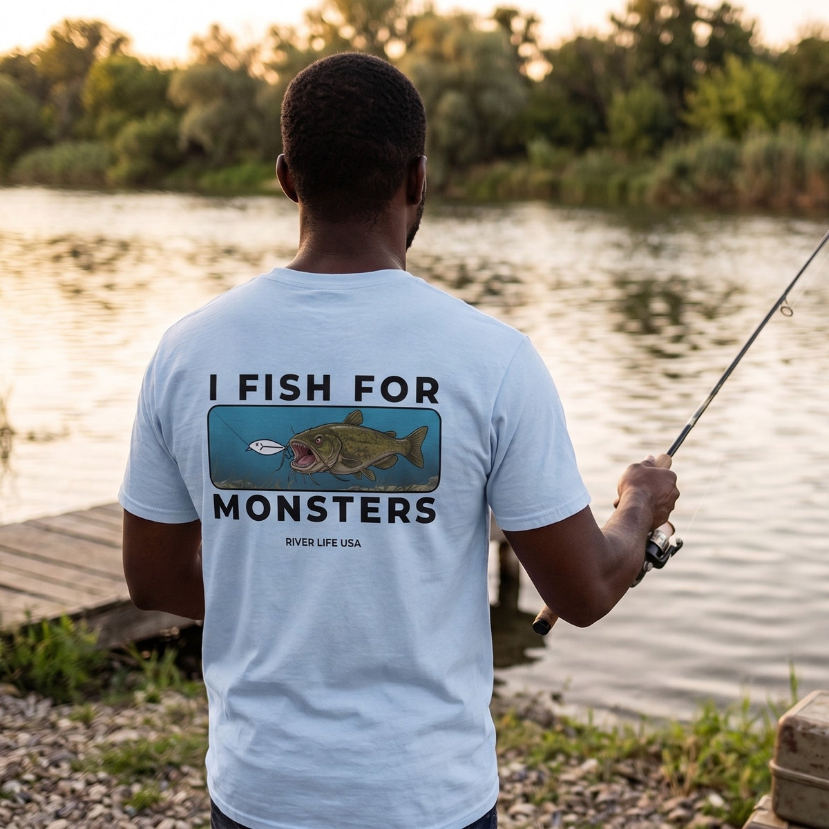 A man stands on a gravel bank by a lake, seen from behind, wearing a white graphic T-shirt that reads 'I FISH FOR MONSTERS' with a catfish illustration and 'River Life USA' below it. He is also wearing a tan cap, dark pants, and rubber boots, with fishing gear and a tan backpack nearby.