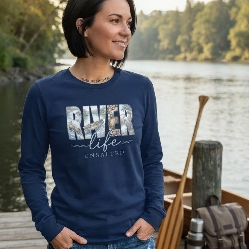 A woman wearing a navy blue long-sleeve crewneck sweatshirt with a 'RIVER life UNSALTED' graphic stands on a wooden dock by a lake, with a canoe and backpack in the background.
